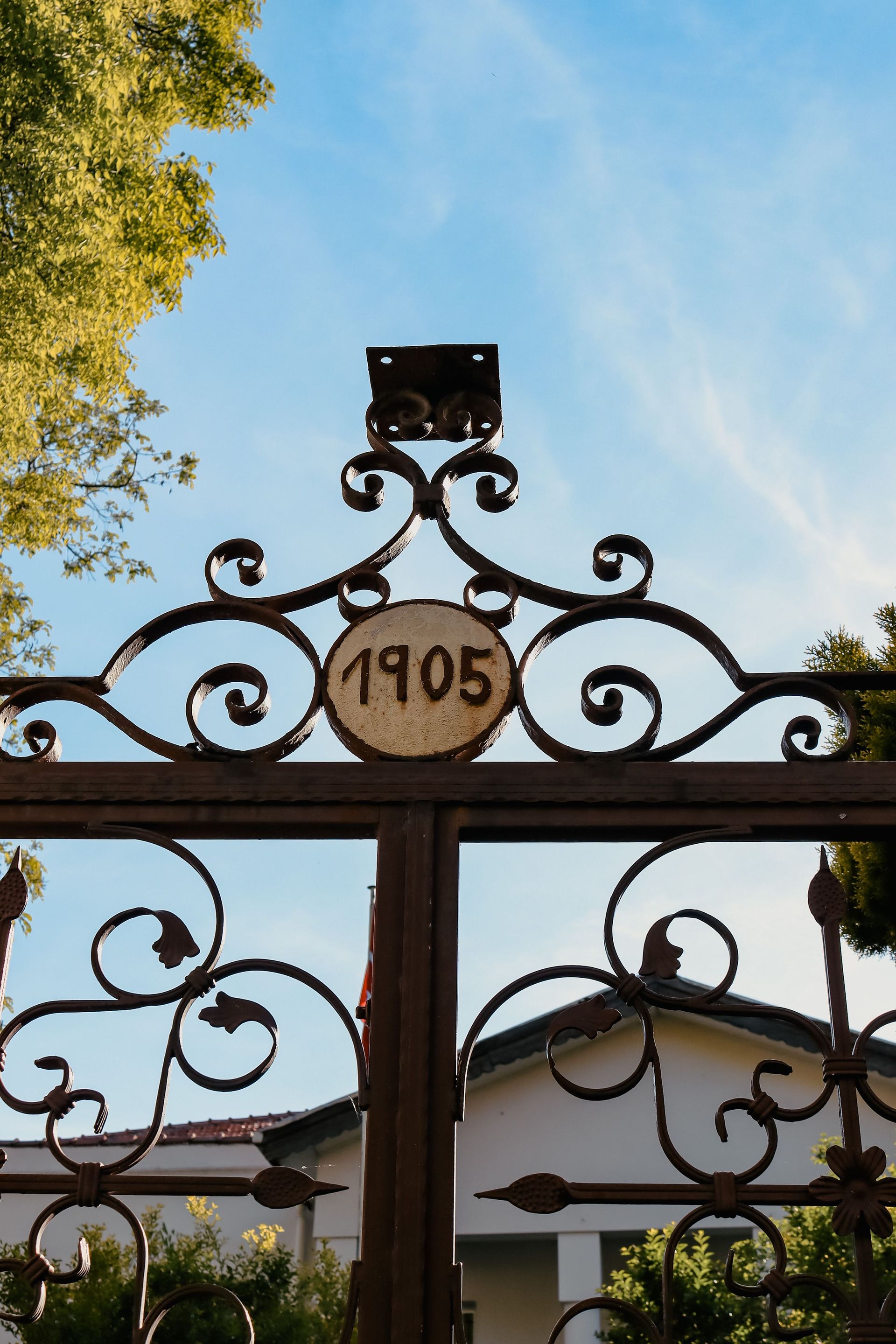 A decorative metal gate with the year 1905 engraved, set against a blue sky, greenery visible.