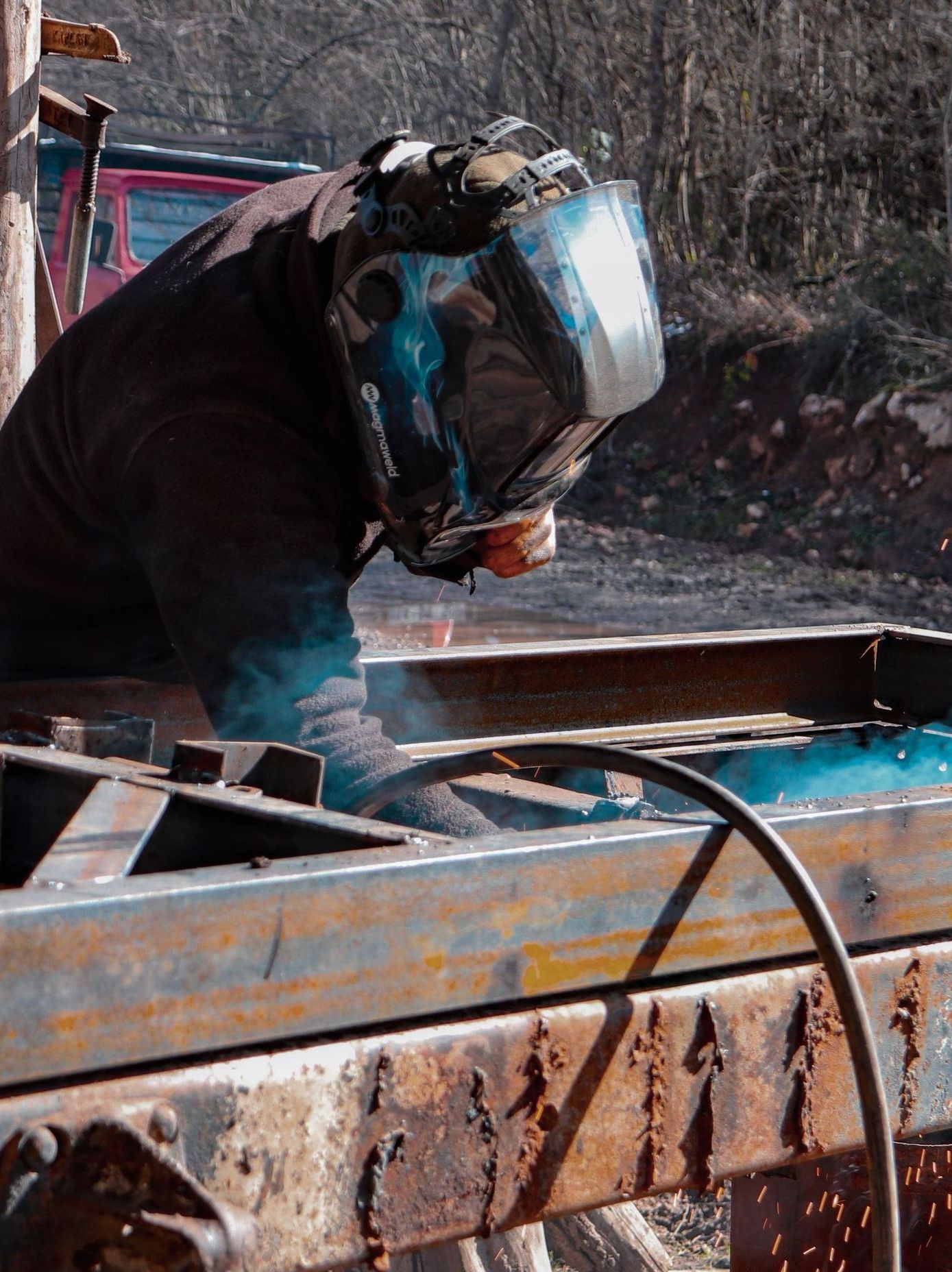 A welder in protective gear works on metal outdoors, creating flying sparks.
