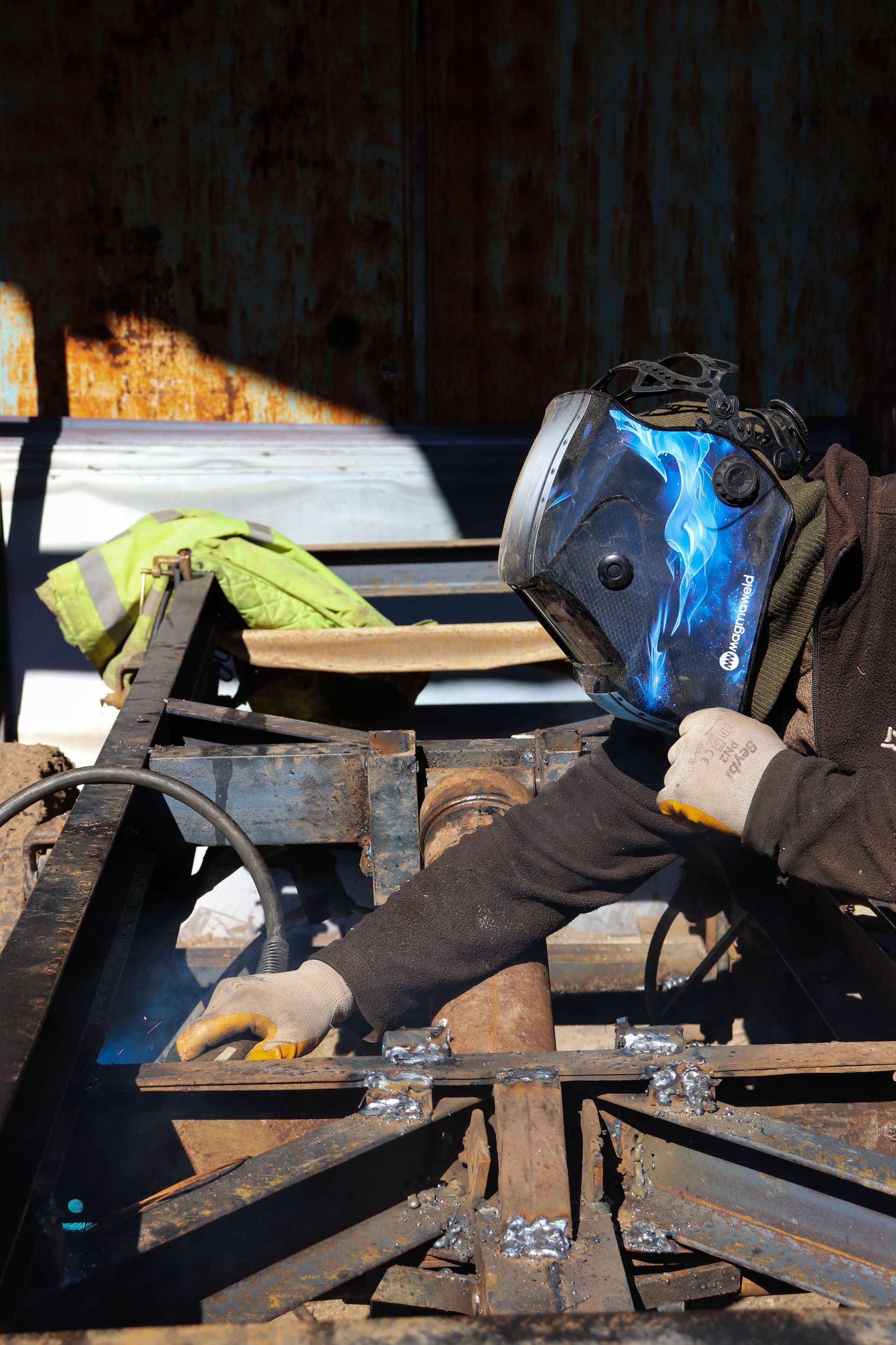 A worker welds metal beams outdoors, showcasing industrial craftsmanship and safety gear.