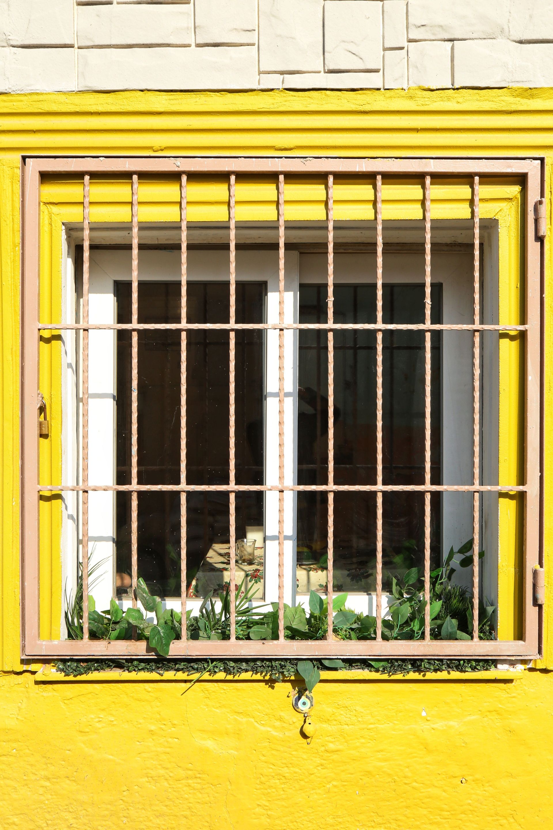 A bright yellow window with bars and greenery, capturing İzmir's unique architectural style.